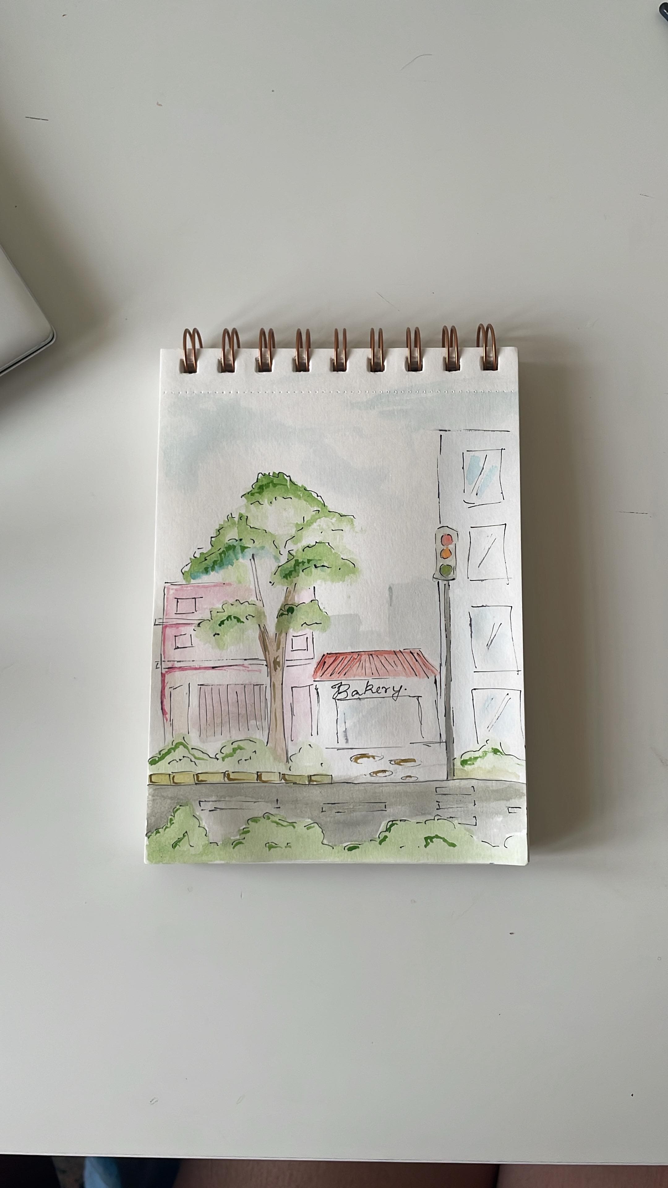 A watercolour street sketch of a bakery beneath a large green tree beside a traffic light and tall pale building.