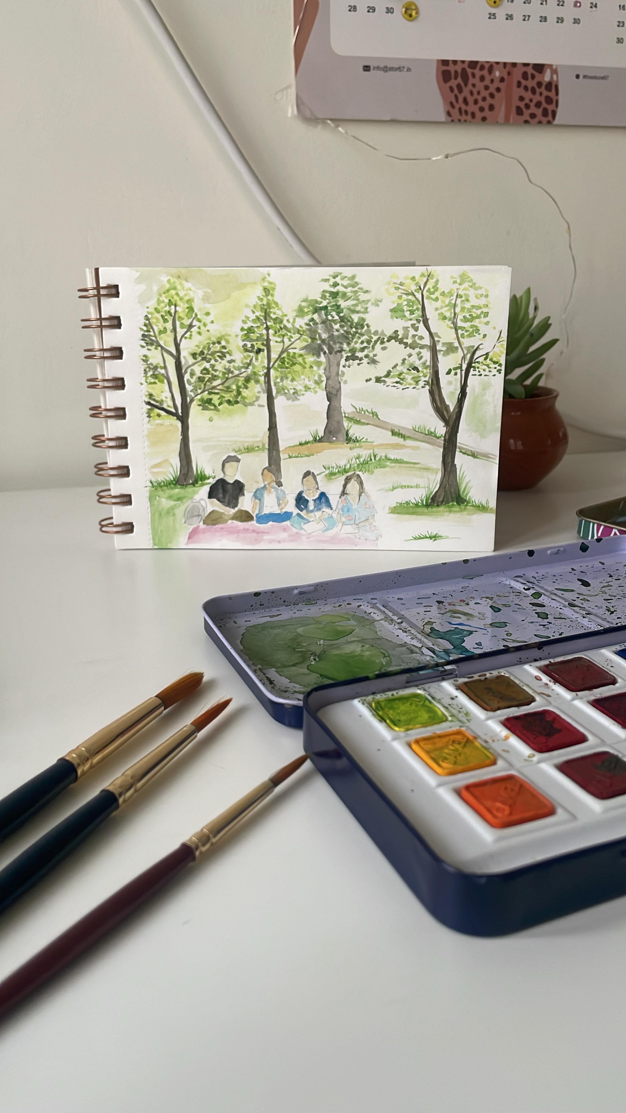 A watercolour of four people picnicking beneath trees, propped behind an open paint tin and several brushes.