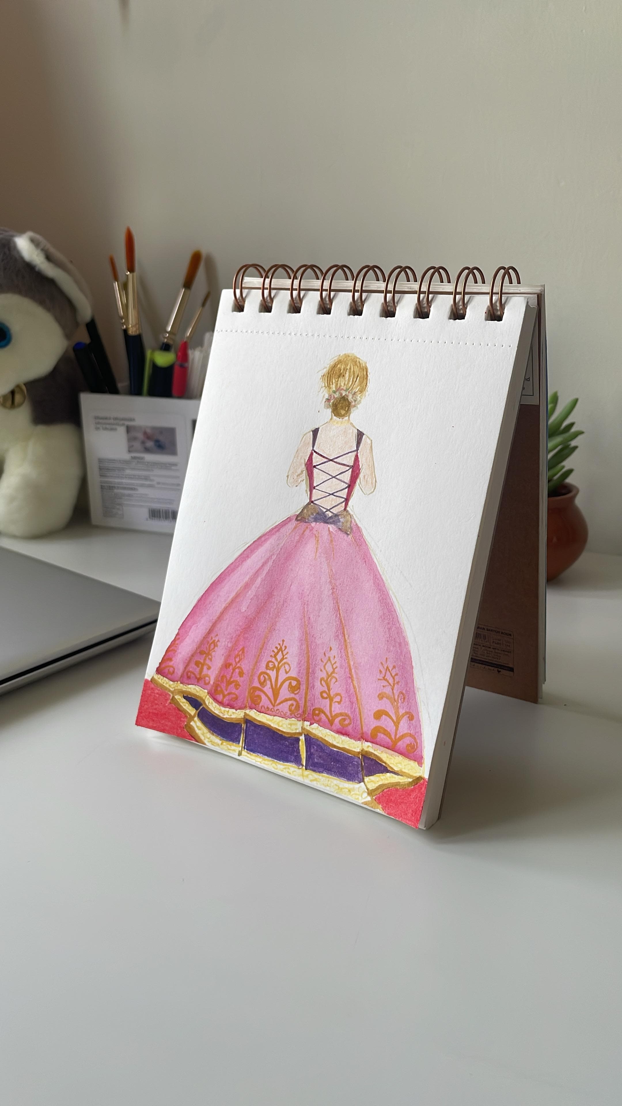 A watercolour figure seen from behind in a wide pink and gold gown, painted in a spiral sketchbook standing on a desk.