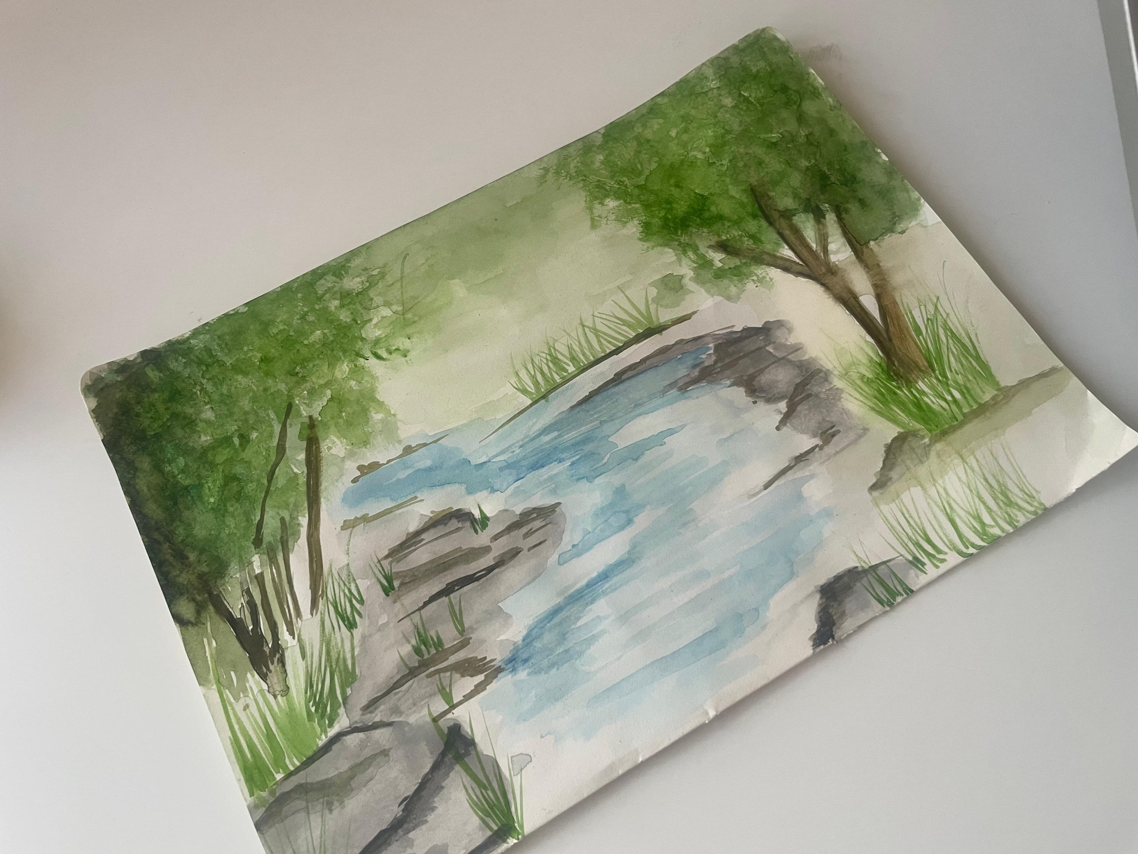 A loose watercolour of a blue stream between green trees and grey rocks, photographed at an angle on a white tabletop.