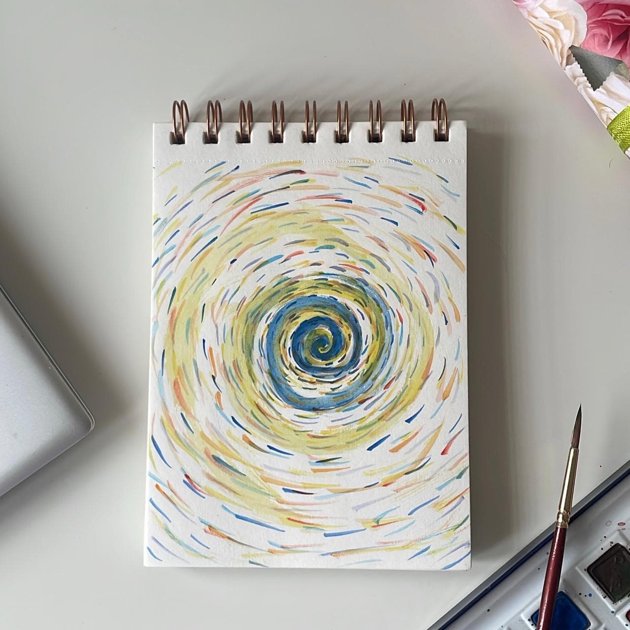 A square studio photograph of a sketchbook page painted with a circular spiral of blue, yellow, green, and coral brush marks.