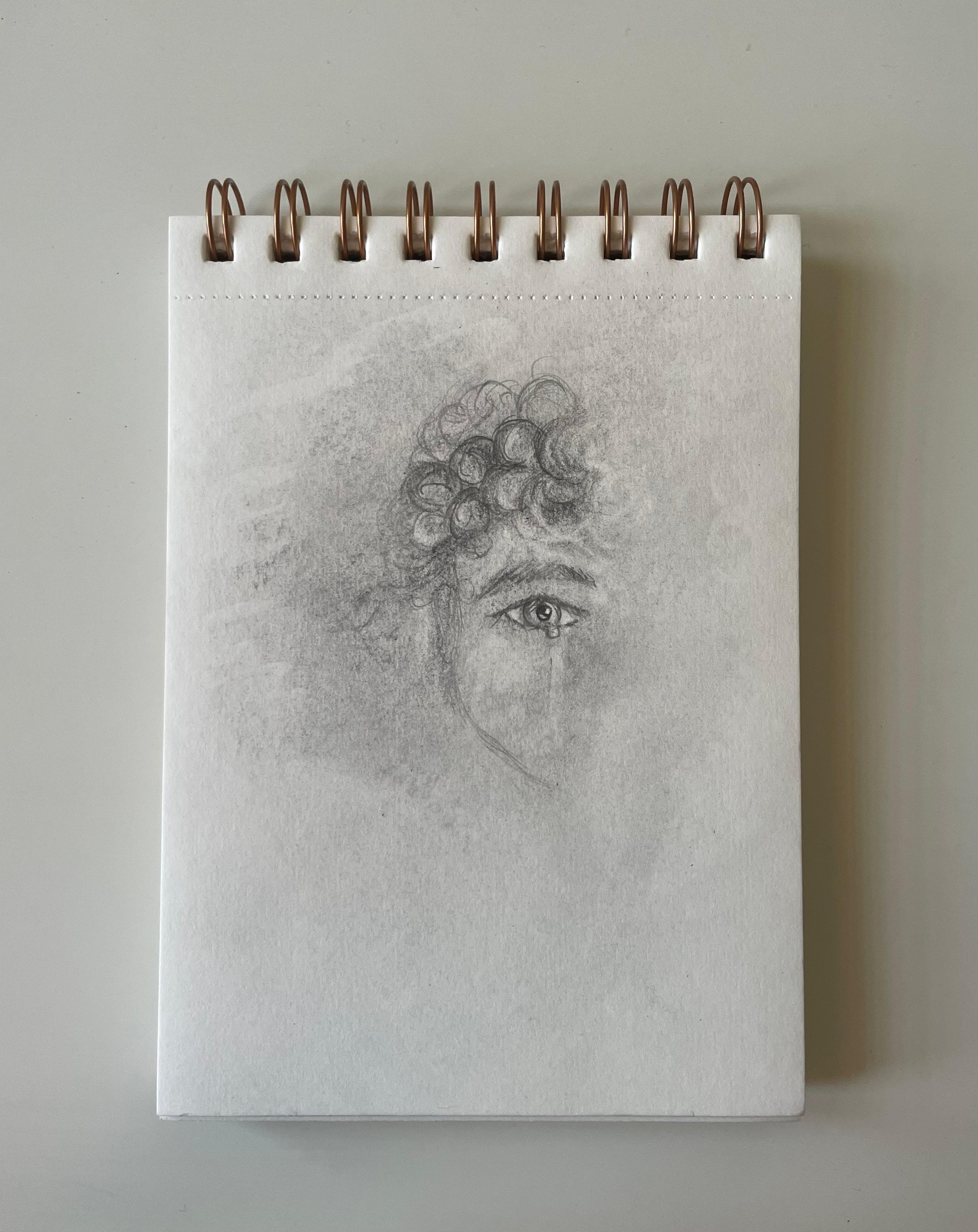A graphite portrait fragment showing one detailed eye with a tear beneath curls, drawn on a vertical spiral sketchbook page.