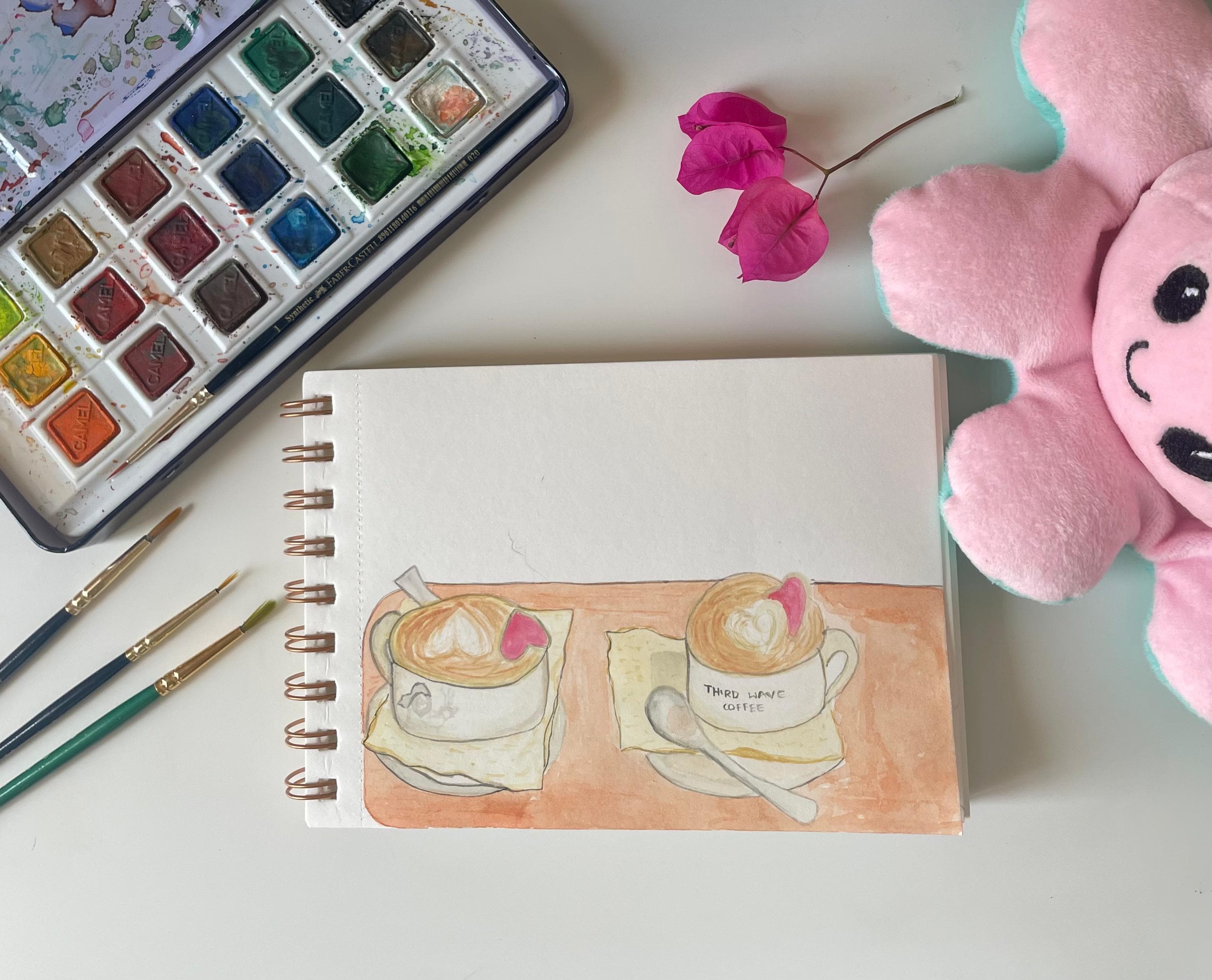 A watercolour of two coffees on a sketchbook page, photographed with a paint box, brushes, magenta flowers, and a pink plush toy.